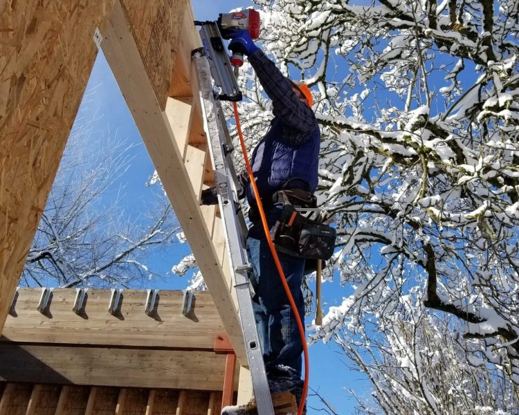 A construction working on a ladder framing an ADU with a tree covered in snow behind him.